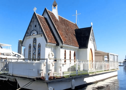 A floating chapel with a brown exterior docked at a marina, featuring stained glass windows and a steeple.