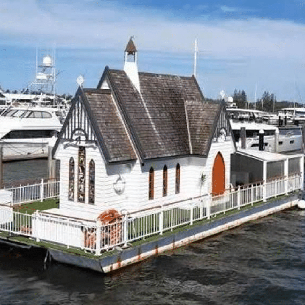 A floating chapel with a brown exterior docked at a marina, featuring stained glass windows and a steeple.