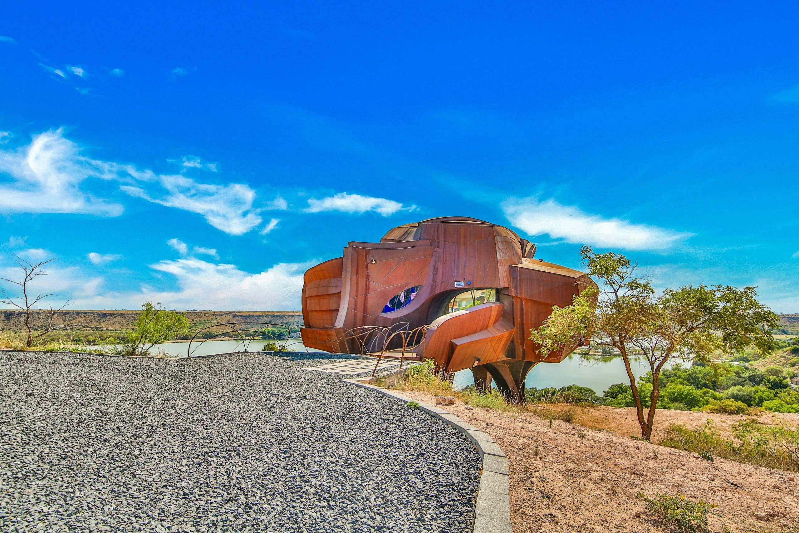 The Robert Bruno Steel House, a rust-colored steel structure on a canyon ridge, with a striking design overlooking a serene lake and scenic landscape.